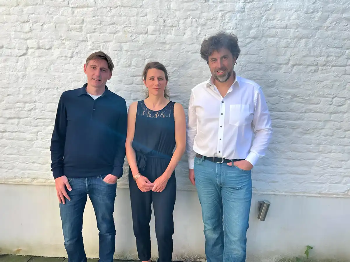Trois personnes debout devant un mur de briques blanches, deux hommes en chemise et jeans, une femme en combinaison bleue.