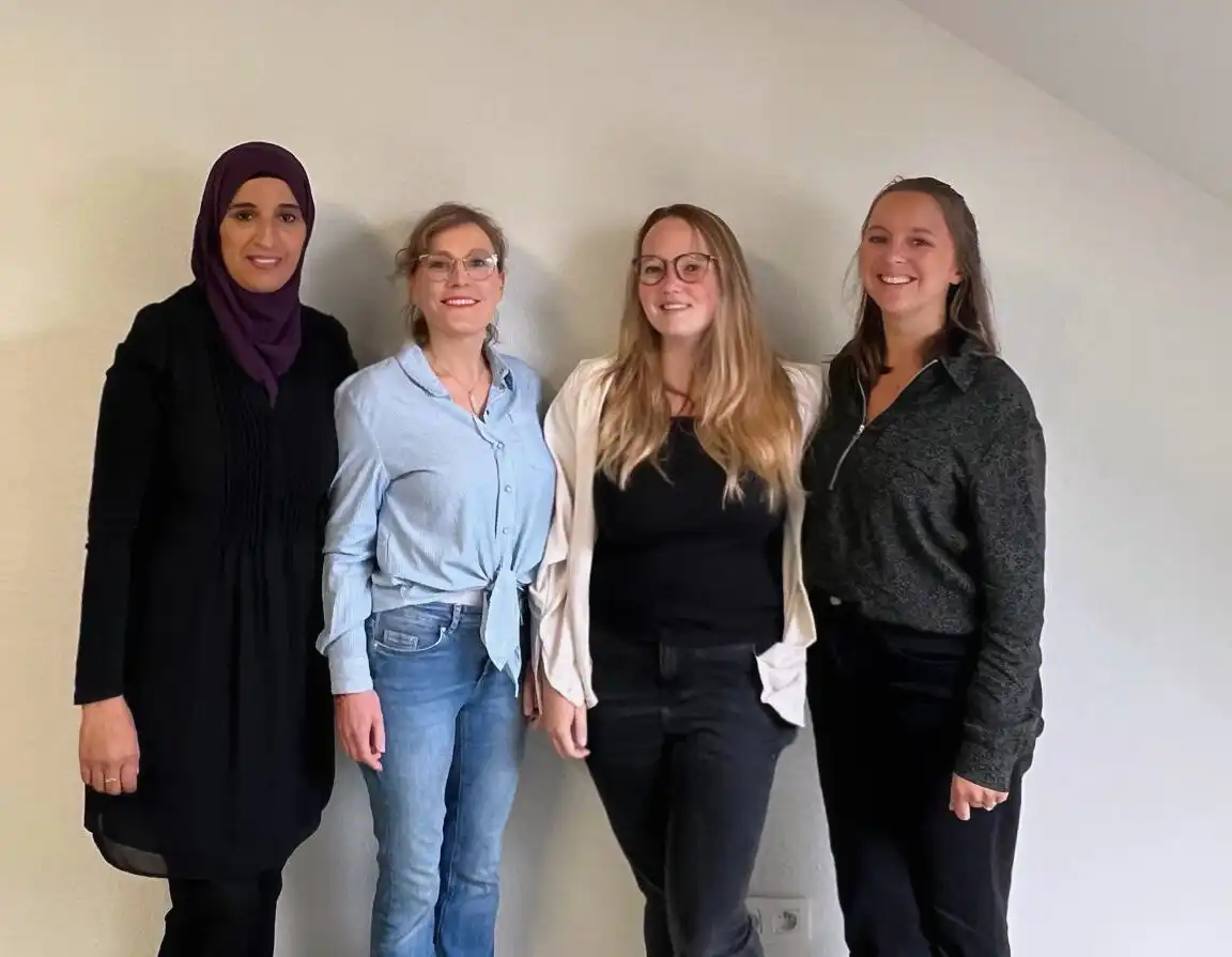Quatre personnes debout contre un mur clair, portant des vêtements variés dans des tons neutres et colorés.