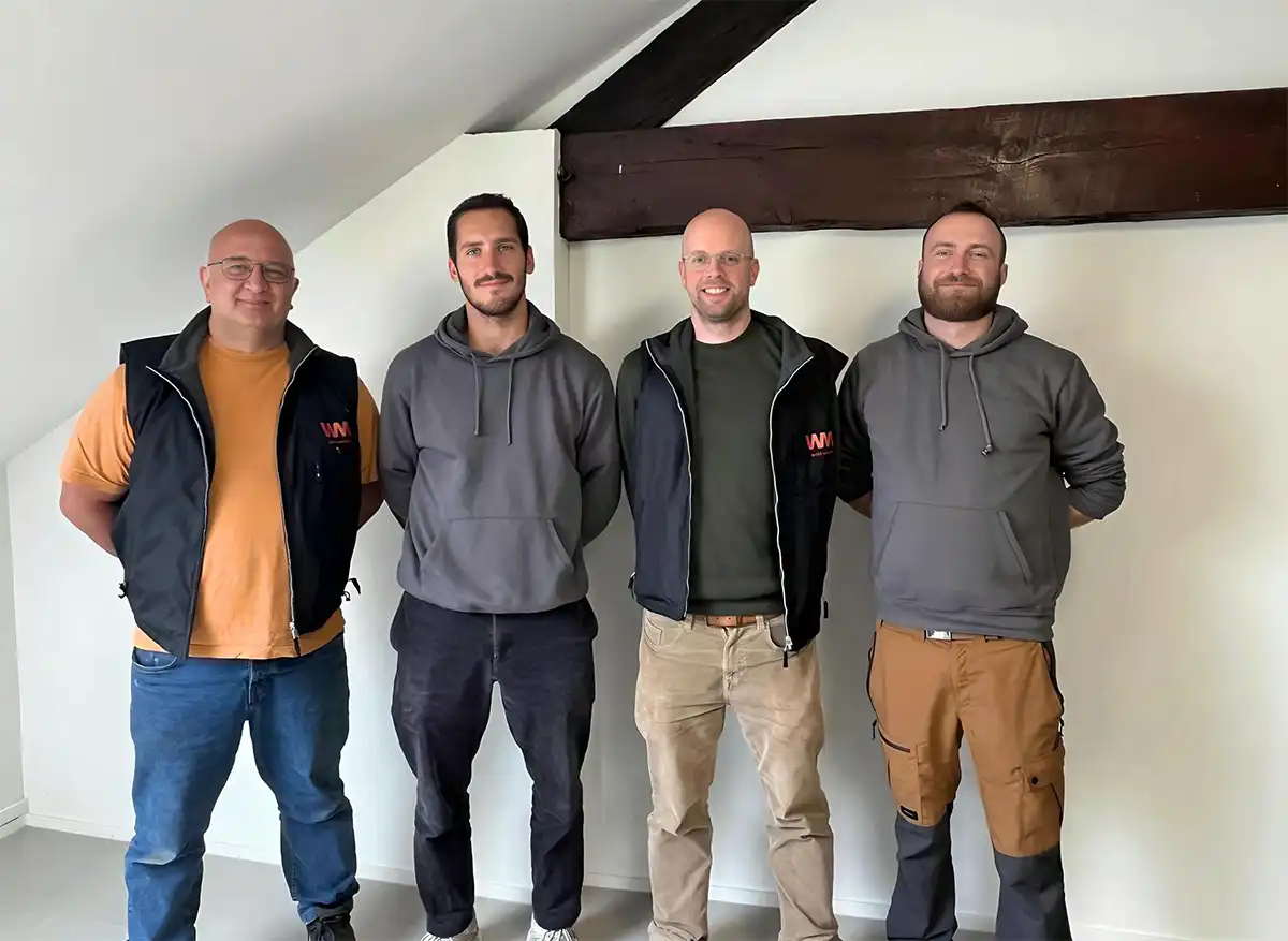 Quatre hommes debout dans une pièce avec un mur blanc et une poutre en bois, portant des vêtements décontractés.