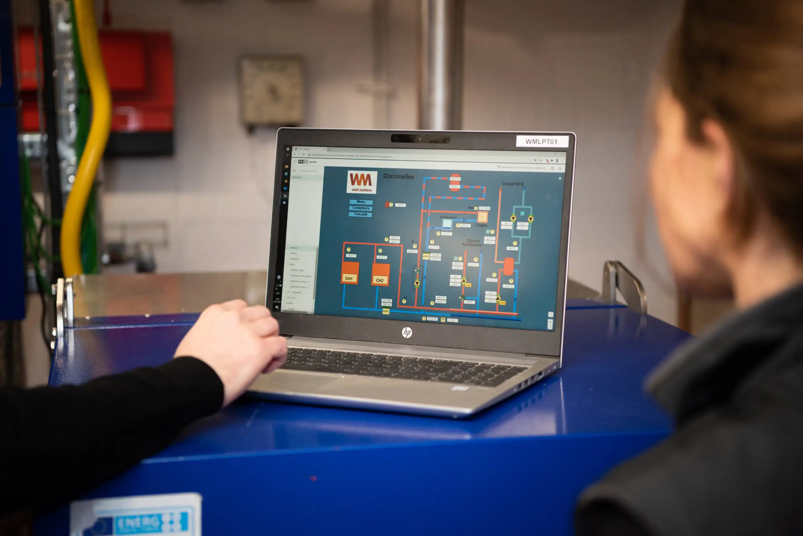 Un ordinateur portable affiche un schéma technique sur son écran, posé sur une surface bleue avec deux personnes à proximité.