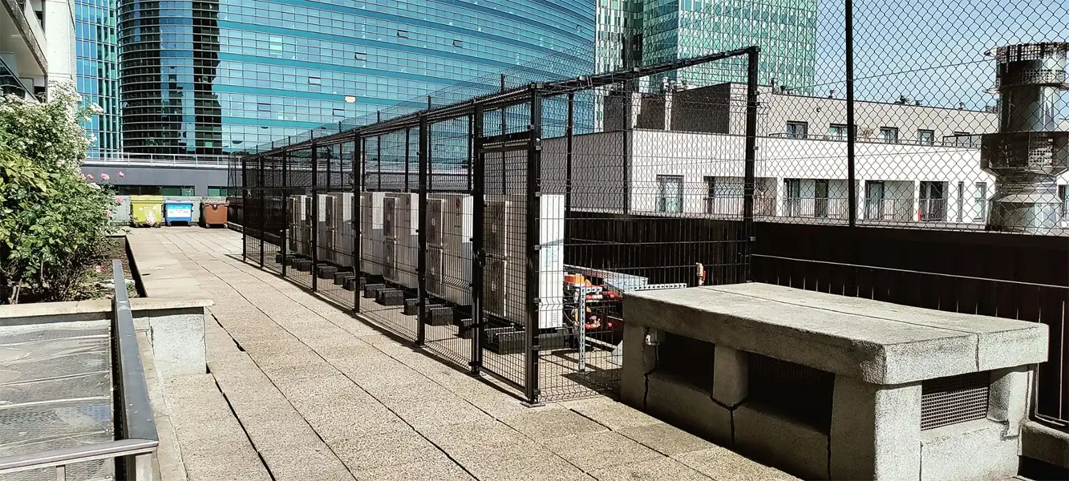 Un alignement de pompes à chaleur derrière une clôture métallique sur une terrasse avec des immeubles modernes en arrière-plan.