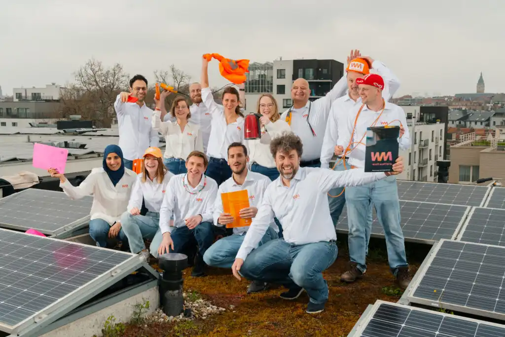 Un groupe de personnes en chemises blanches pose sur un toit avec des panneaux solaires, tenant des objets orange et roses.