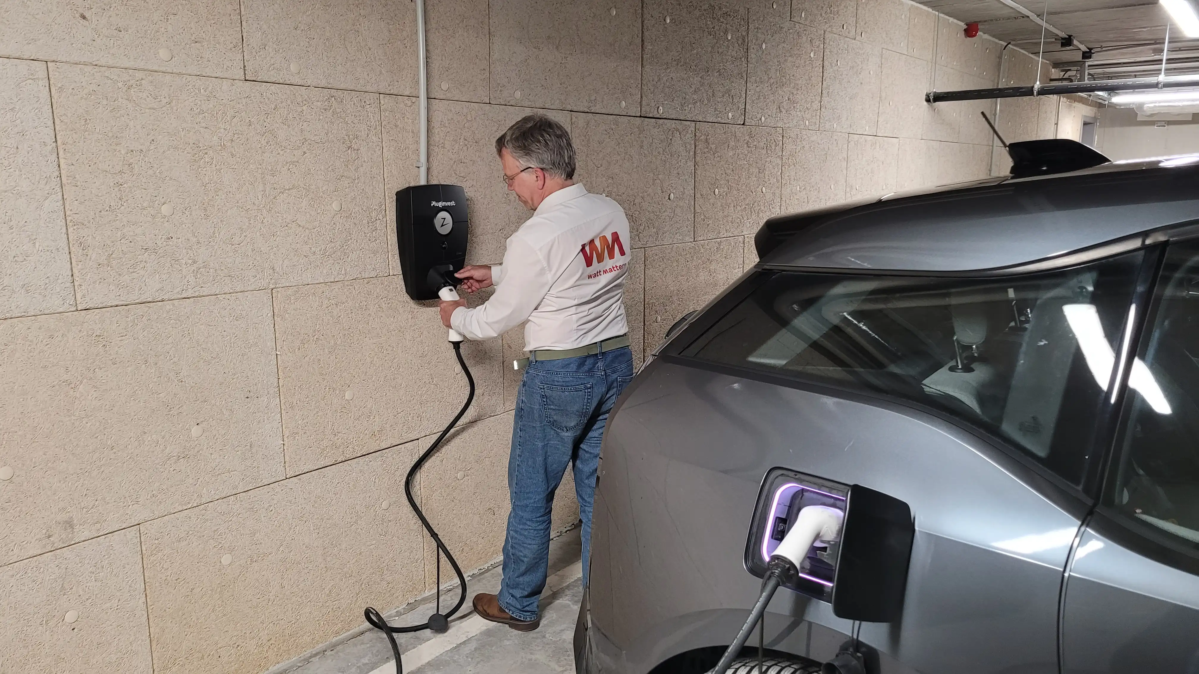 Un homme branche une voiture électrique grise à une borne de recharge murale dans un garage.