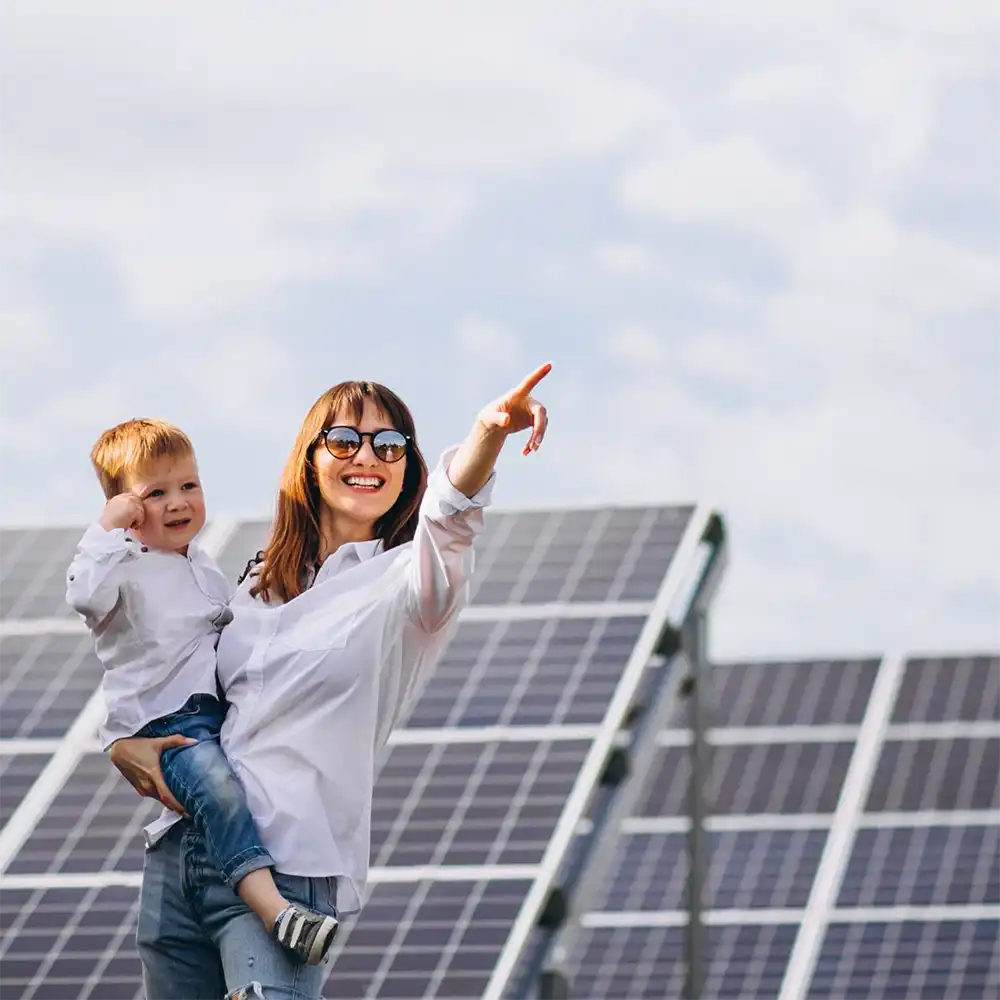 Une femme souriante portant des lunettes de soleil tient un enfant devant des panneaux solaires sous un ciel nuageux.