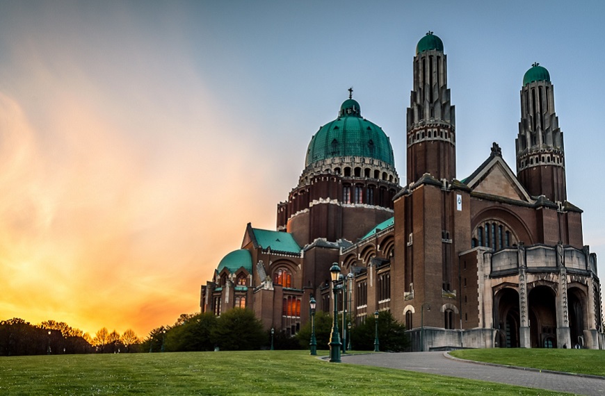 La Basilique de Koekelberg au crépuscule, avec son dôme vert et ses tours, sur un fond de ciel orangé.