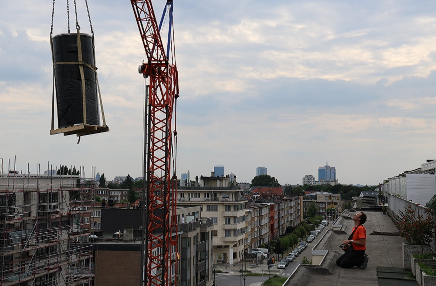 Un homme en gilet orange contrôle une grue rouge soulevant un objet cylindrique sur un toit, avec une vue urbaine en arrière-plan.