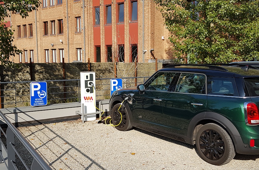 Voiture verte électrique en charge sur une borne dans un parking réservé, devant un bâtiment en briques.