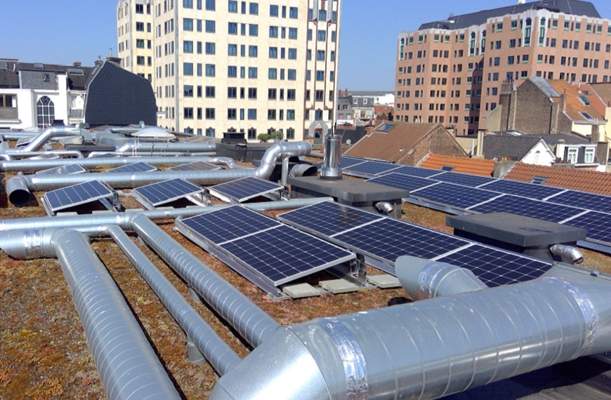 Panneaux solaires et conduits métalliques sur un toit, entourés de bâtiments en briques et en béton sous un ciel bleu.