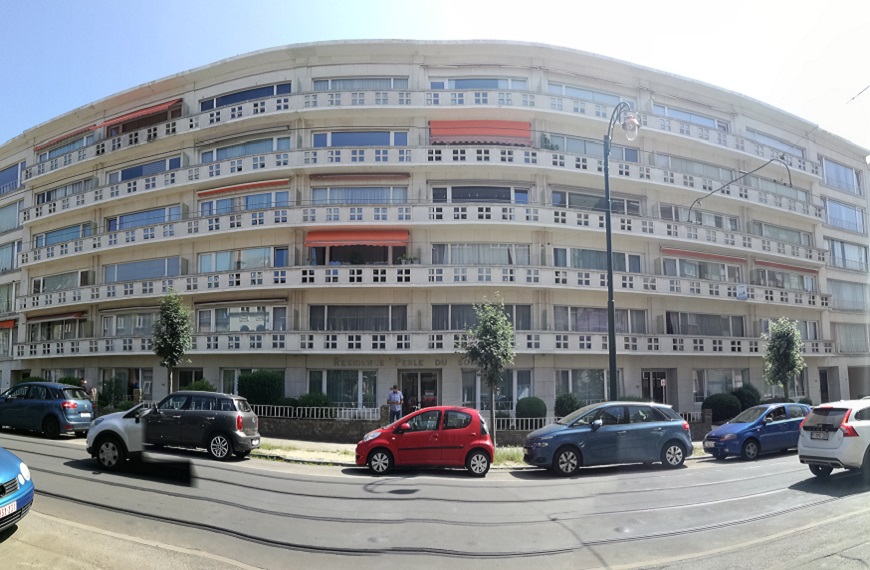Immeuble résidentiel moderne avec balcons vitrés et stores orange, situé le long d'une rue avec plusieurs voitures garées.
