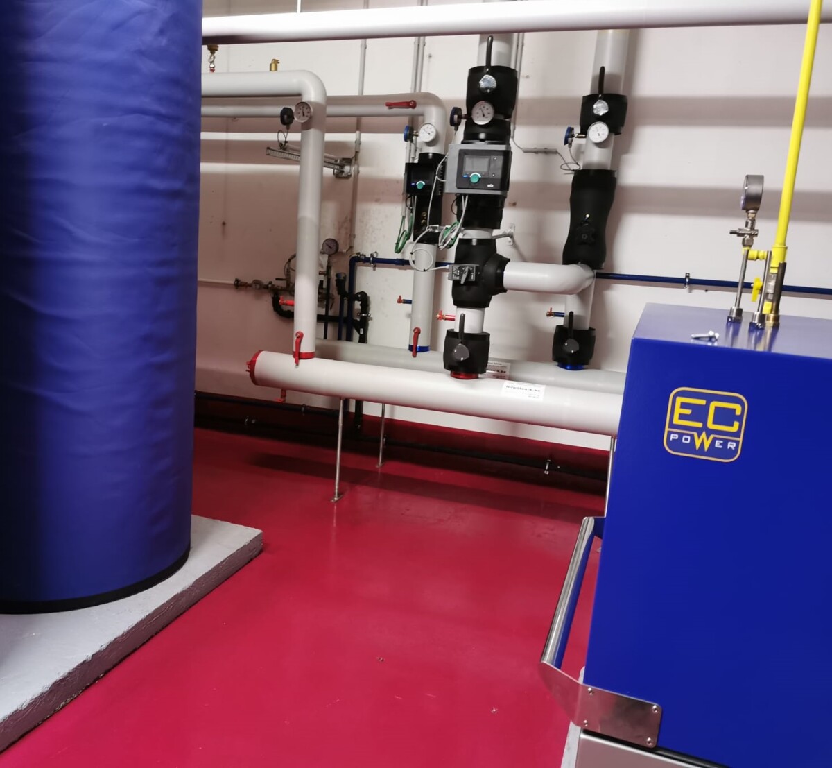 Installation de tuyauterie industrielle avec réservoir bleu et unité EC Power sur sol rouge dans une salle technique.