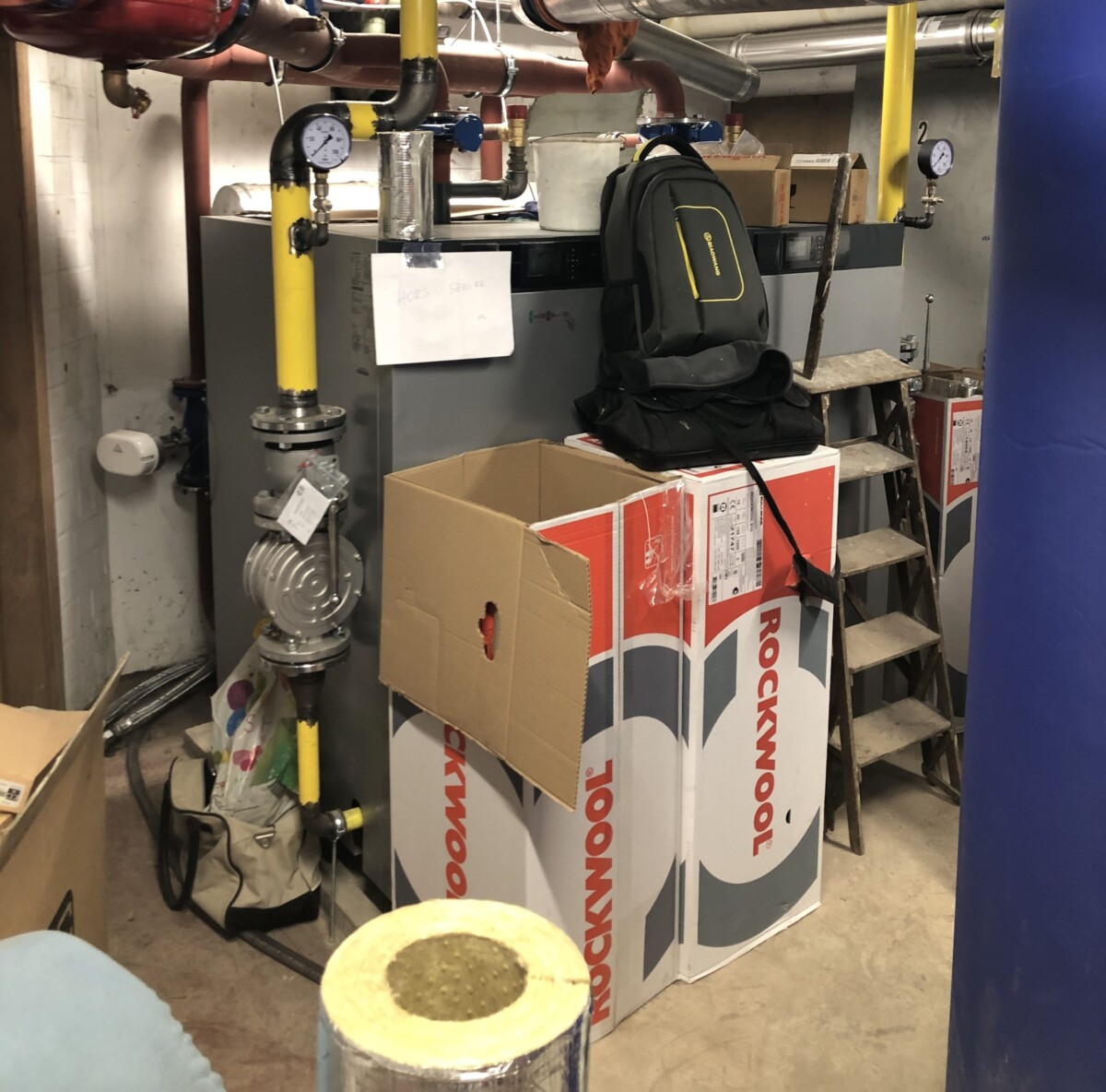 Un local technique avec des tuyaux colorés, des boîtes Rockwool, un carton ouvert, un sac à dos noir et une échelle en bois.
