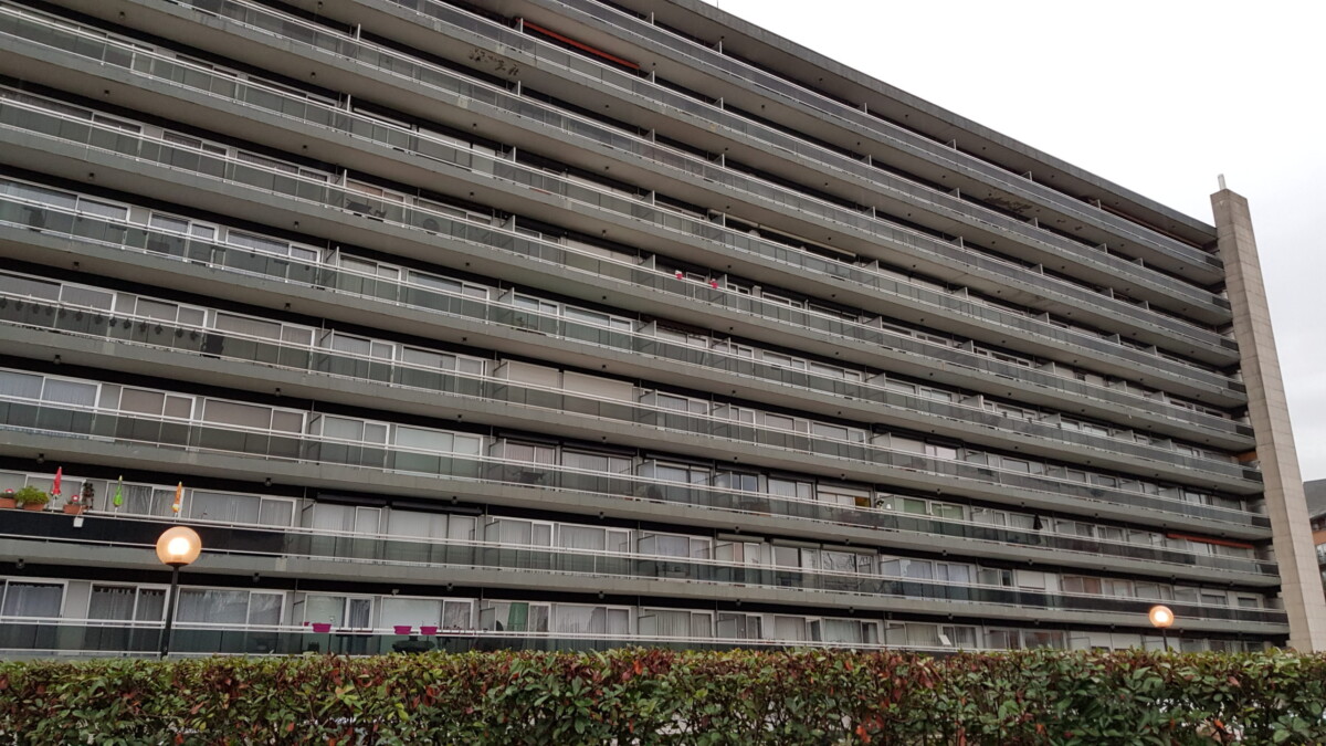 Un grand immeuble résidentiel gris avec des balcons alignés, entouré de végétation et de lampadaires allumés au premier plan.