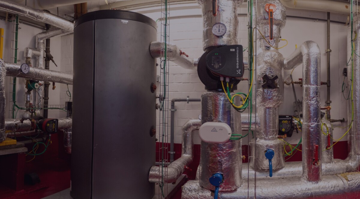 Installation de plomberie industrielle avec réservoir gris et tuyaux isolés en métal dans une salle technique.