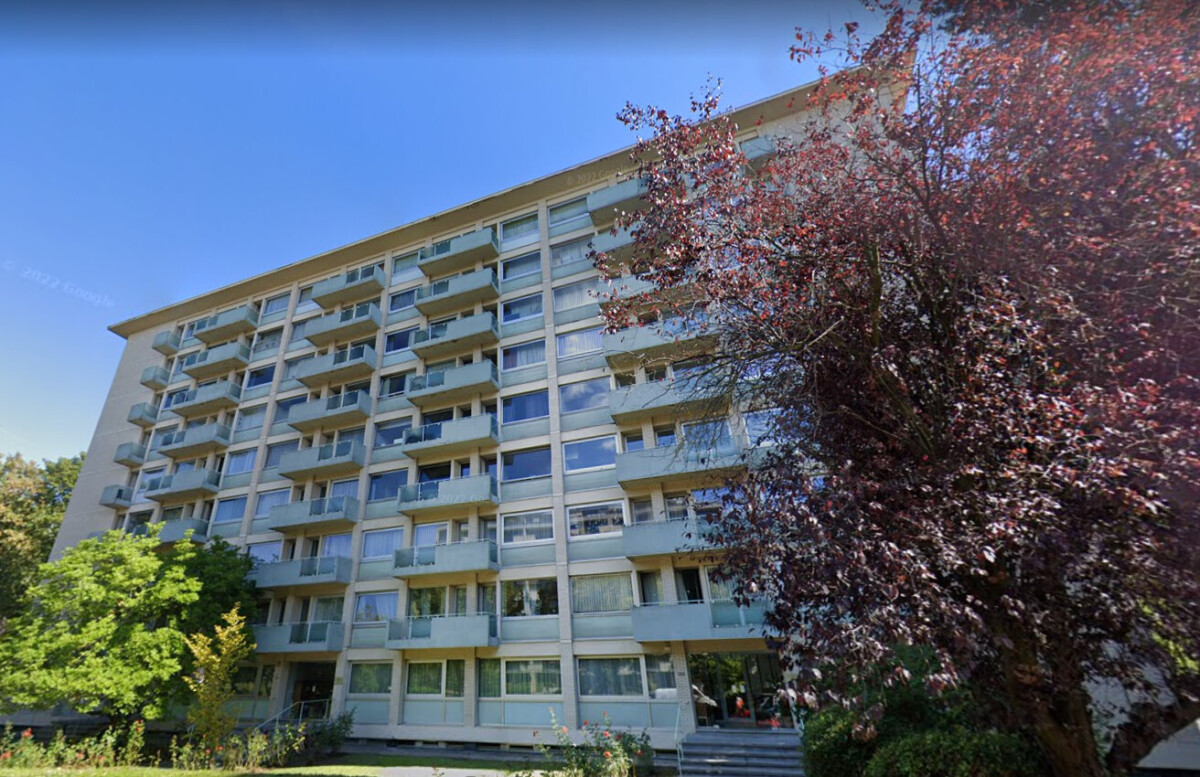 Immeuble résidentiel beige à plusieurs étages avec balcons, entouré d'arbres feuillus sous un ciel bleu.