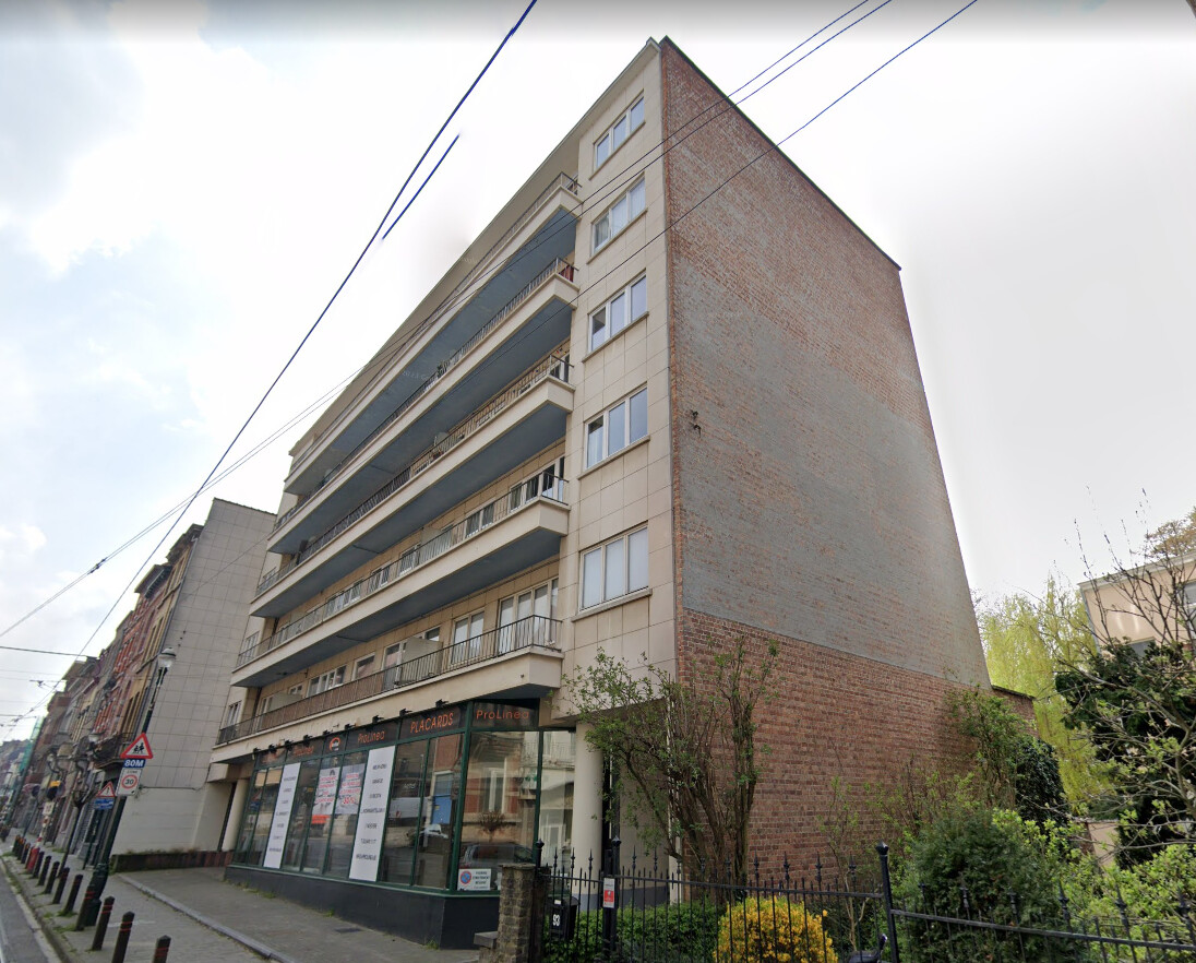 Immeuble résidentiel en briques rouges et beige avec balcons, situé au coin d'une rue bordée de végétation.