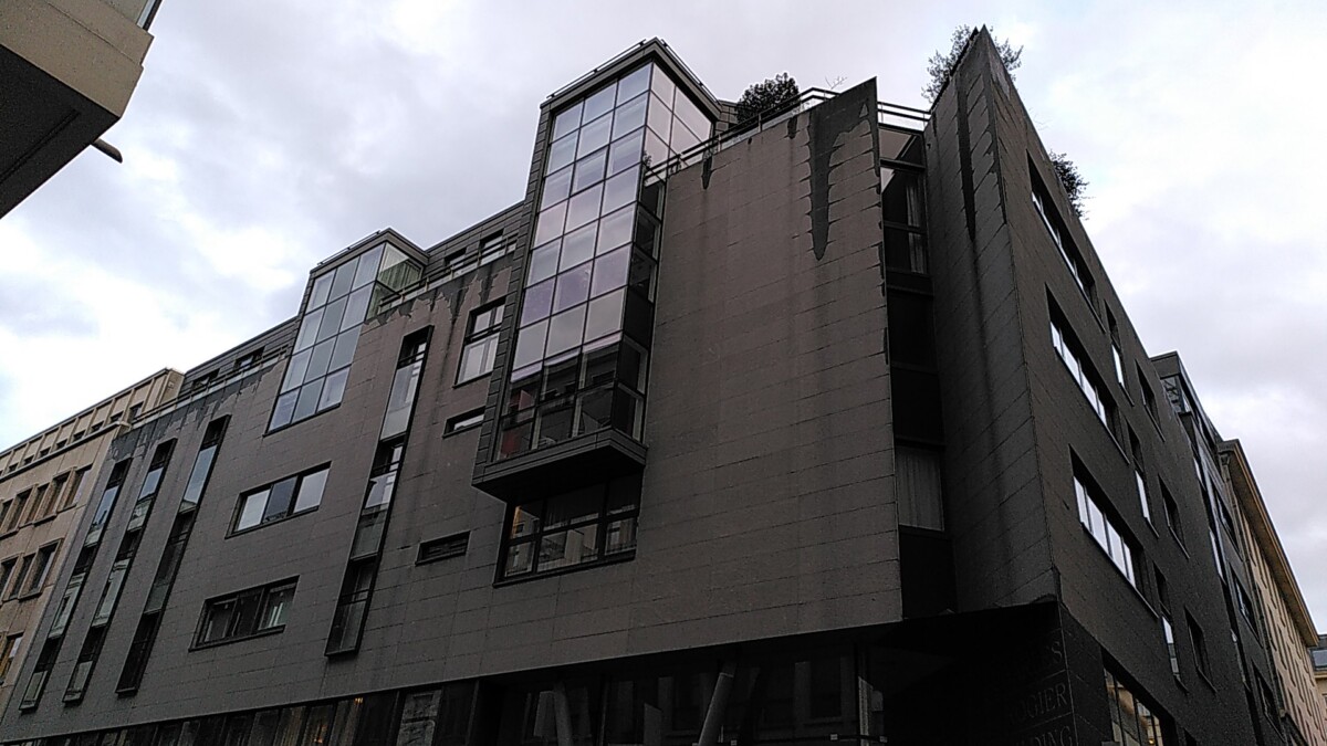 Bâtiment moderne avec façade en verre et métal, nommé Charles Rogier, sous un ciel nuageux.