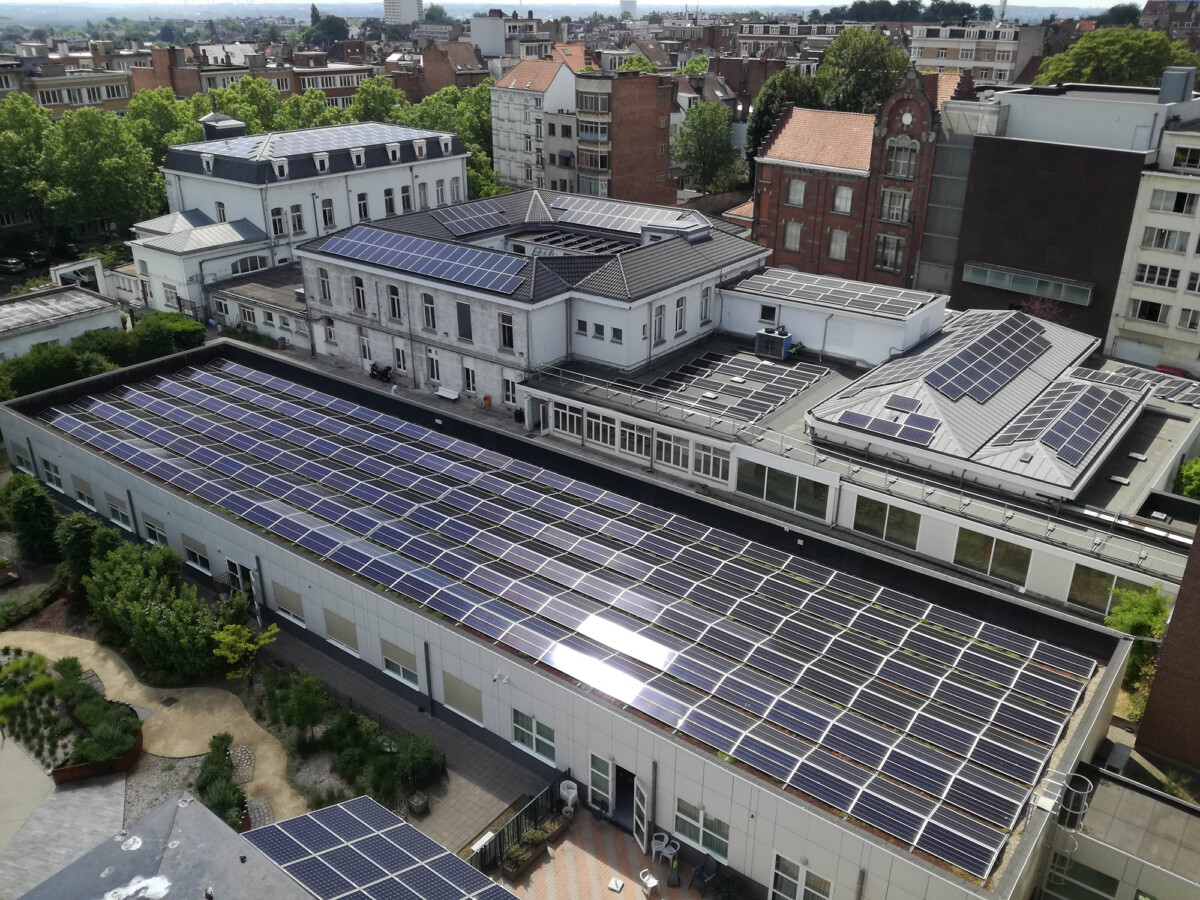 Vue aérienne de bâtiments avec toits équipés de panneaux solaires, entourés de végétation et d'autres immeubles en arrière-plan.