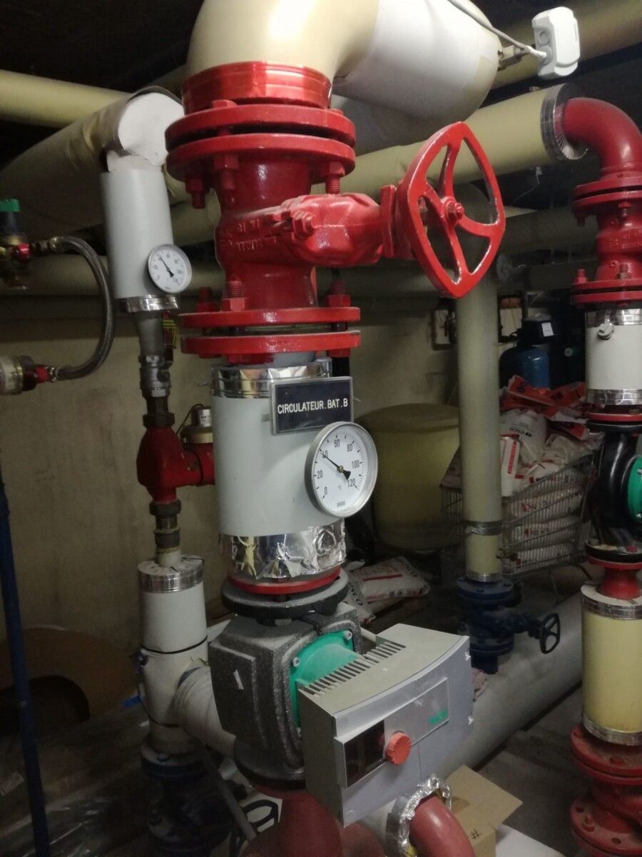 Des tuyaux industriels rouges et beiges avec des manomètres et vannes rouges dans une salle technique.
