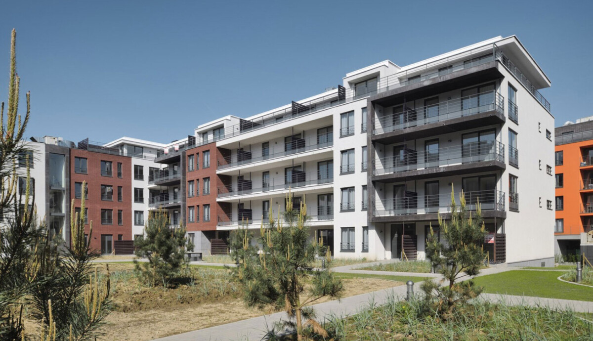 Immeuble résidentiel moderne avec façades blanches et rouges, entouré de jardinets et d'allées pavées, sous un ciel bleu.