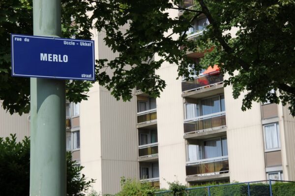 Panneau de rue "Merlo" à Uccle devant un immeuble beige avec balcons, partiellement caché par des arbres.