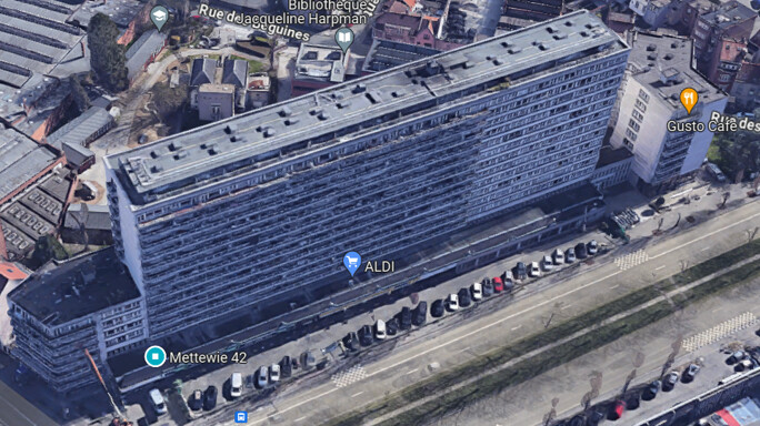 Vue aérienne d'un grand immeuble résidentiel rectangulaire à Mettewie 46, avec un parking et des voitures alignées devant.