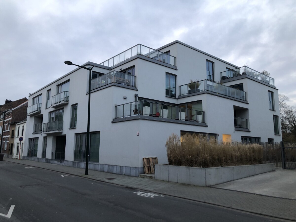 Immeuble moderne blanc avec balcons vitrés et plantes, situé à l'angle d'une rue pavée sous un ciel nuageux.