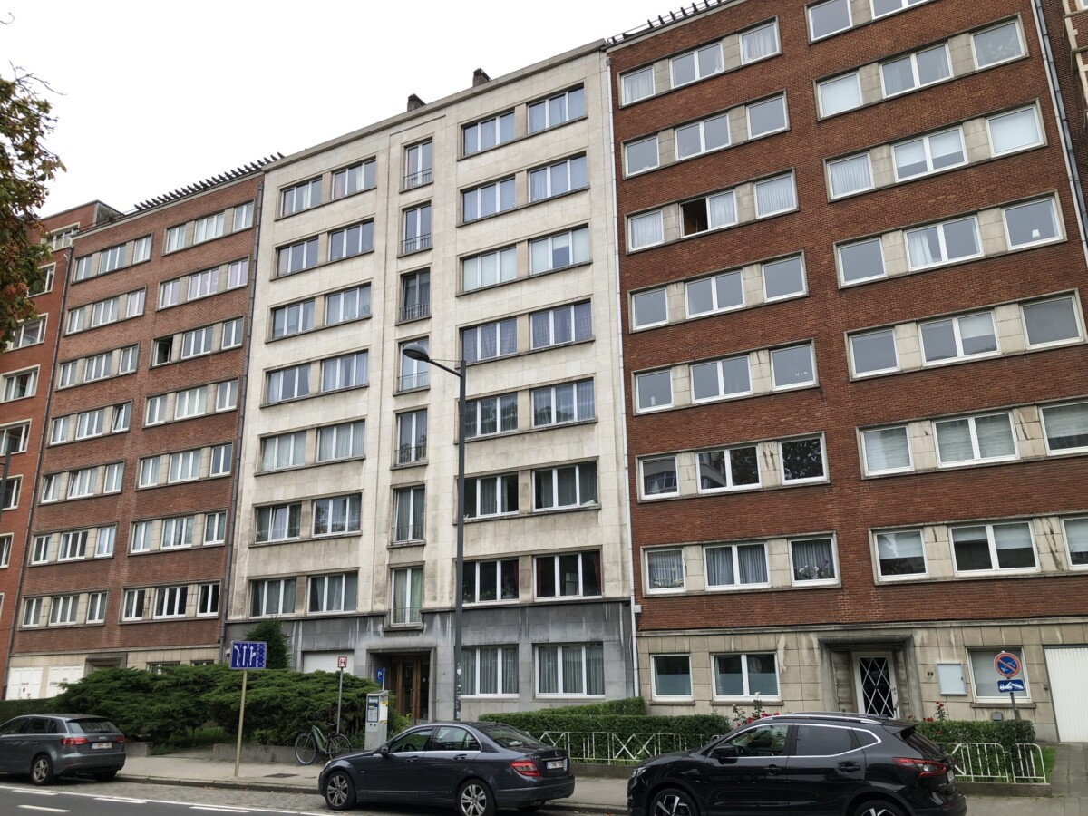 Immeuble résidentiel en briques rouges et blanches avec plusieurs étages, voitures garées devant sur une rue arborée.