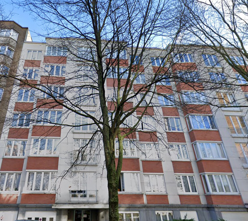 Immeuble résidentiel en briques rouges et blanches avec de grandes fenêtres, devant un arbre dénudé sous un ciel bleu.
