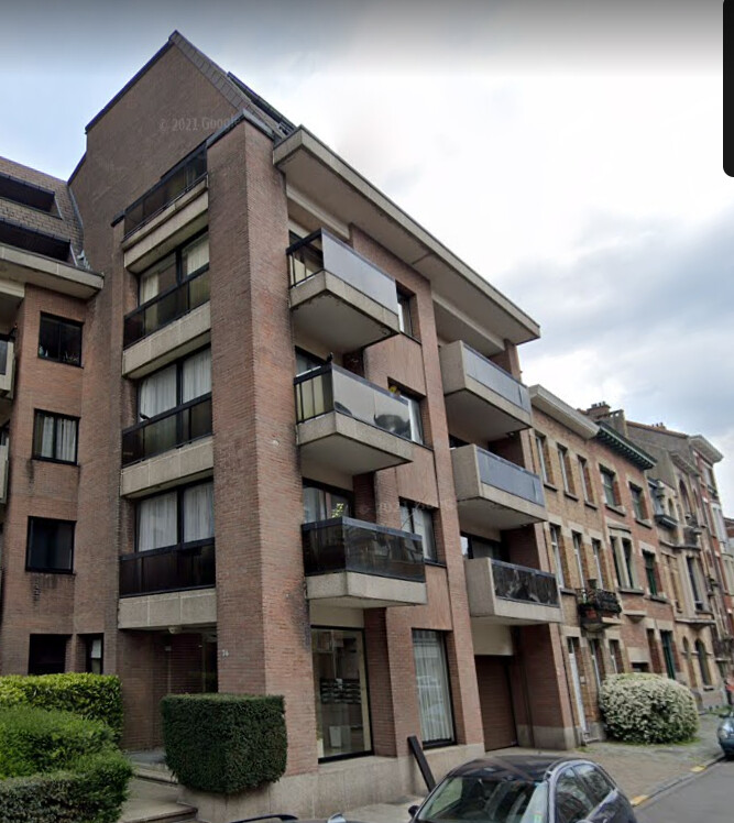 Immeuble résidentiel en briques rouges avec balcons vitrés, situé dans une rue bordée de voitures et de végétation.