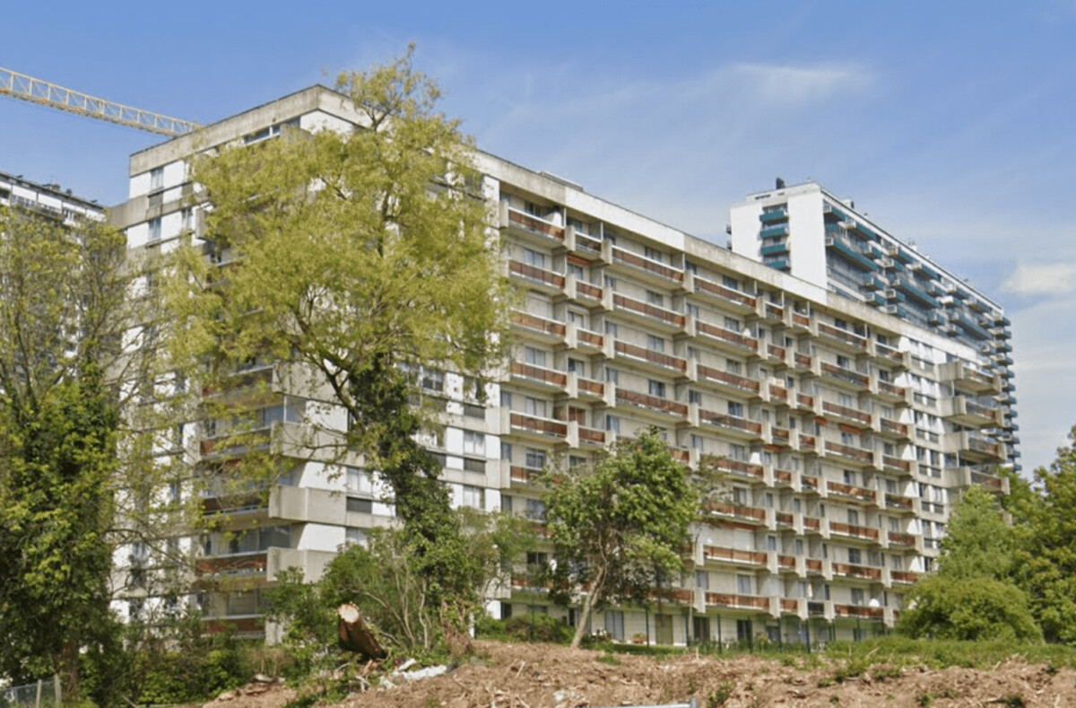 Immeuble résidentiel moderne entouré d'arbres avec un ciel bleu en arrière-plan, grue visible à gauche.