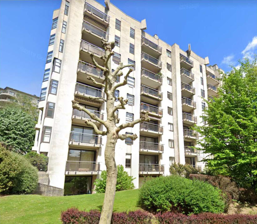 Immeuble résidentiel beige avec balcons, entouré d'arbres et de buissons verts sous un ciel bleu.