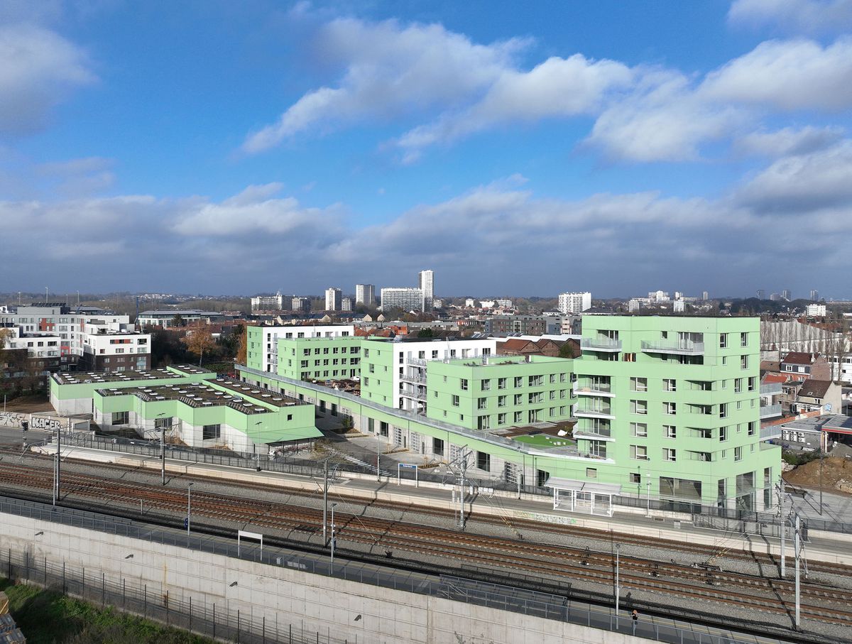 Bâtiments résidentiels verts de Novacity à côté de voies ferrées, sous un ciel partiellement nuageux, avec une vue urbaine en arrière-plan.