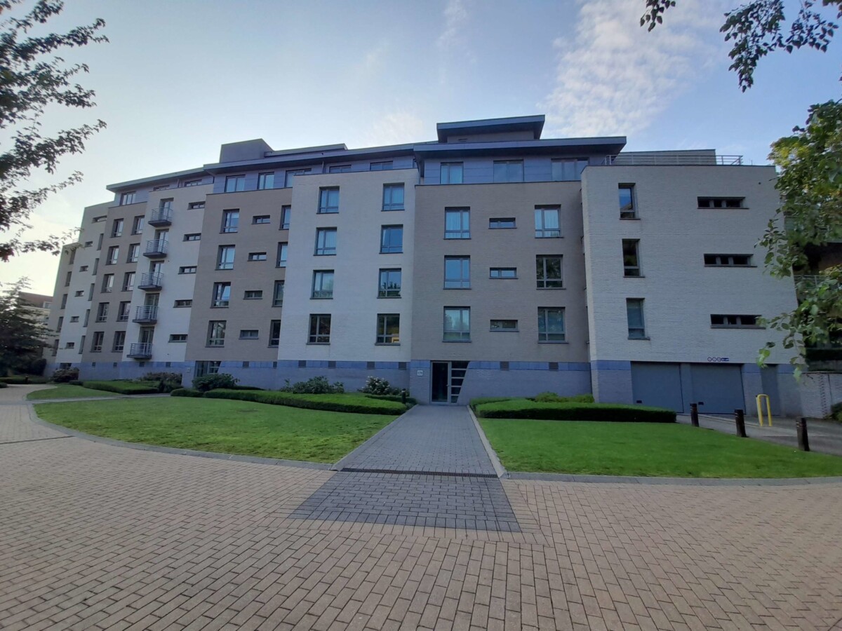 Immeuble résidentiel moderne de cinq étages avec façades en briques claires, entouré d'un jardin et d'une allée pavée.