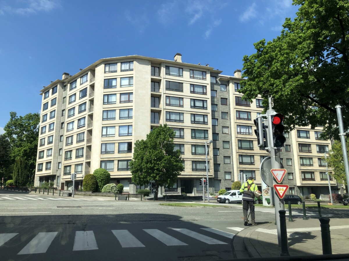 Immeuble résidentiel beige de plusieurs étages avec des fenêtres multiples, situé à un carrefour avec feu de signalisation rouge et panneaux de signalisation.